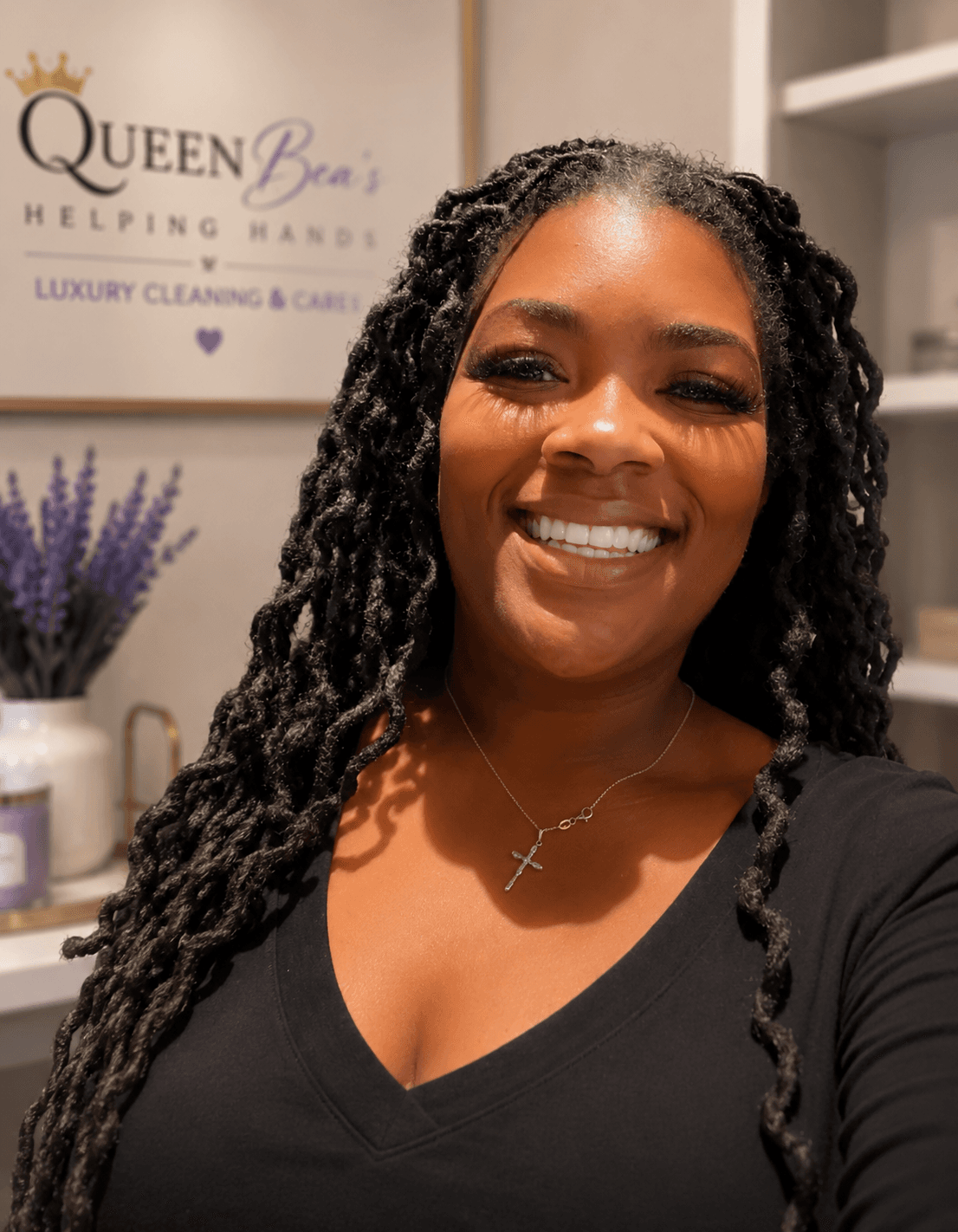 Smiling woman with long dark twists and cross necklace before a Queen Bea's sign.